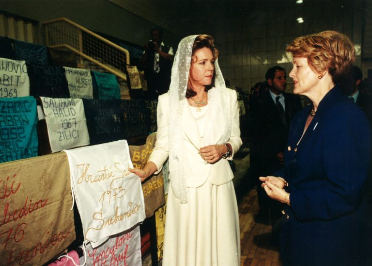 Queen Noor in mourning white, holds a cloth embroidered by a woman with the name of a murdered brother, son, father, or husband. The stadium was blanketed with thousands of these reminders.
